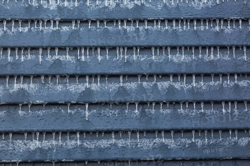 Close-up of exterior garden shed wall made of grey clapboard siding and covered with ice and hanging icicles after freezing rain in late winter, Quebec, Canada