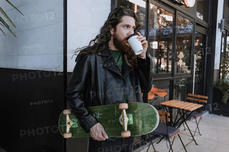 A skateboarder in a black leather jacket takes a sip of coffee while holding a skateboard in an urban setting. Outdoor relaxation and street style