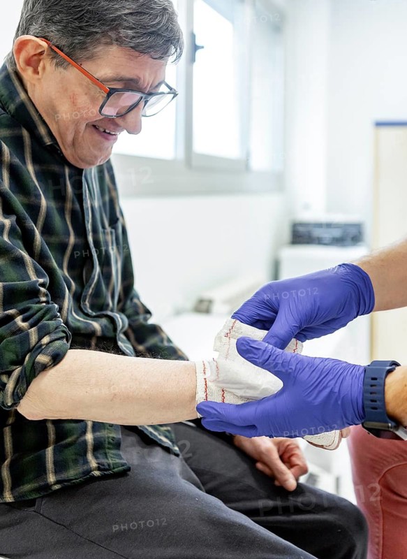 A healthcare professional tends to a patient's arm in a mental health residence. The caregiver wears gloves, highlighting support and care in a therapeutic setting