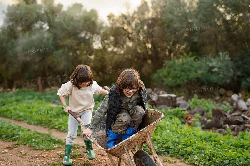 Two children engage in a joyful wheelbarrow ride in a rustic outdoor setting. Surrounded by greenery, they exude carefree happiness and childhood delight