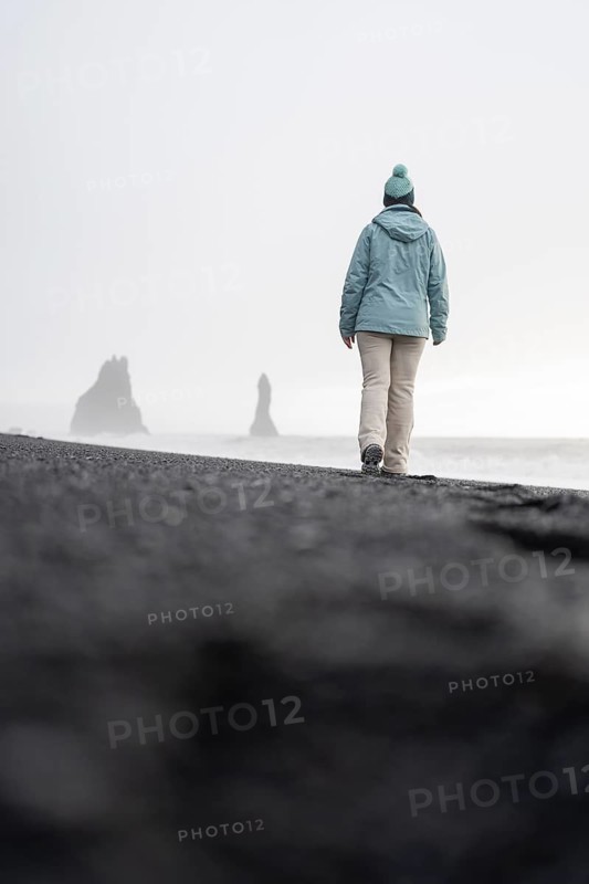 Back view of an unrecognizable of a solitary woman wanders towards the famous Reynisdrangar sea stacks on the misty black sands of Reynisfjara Beach in Iceland, capturing a tranquil yet eerie winter scene