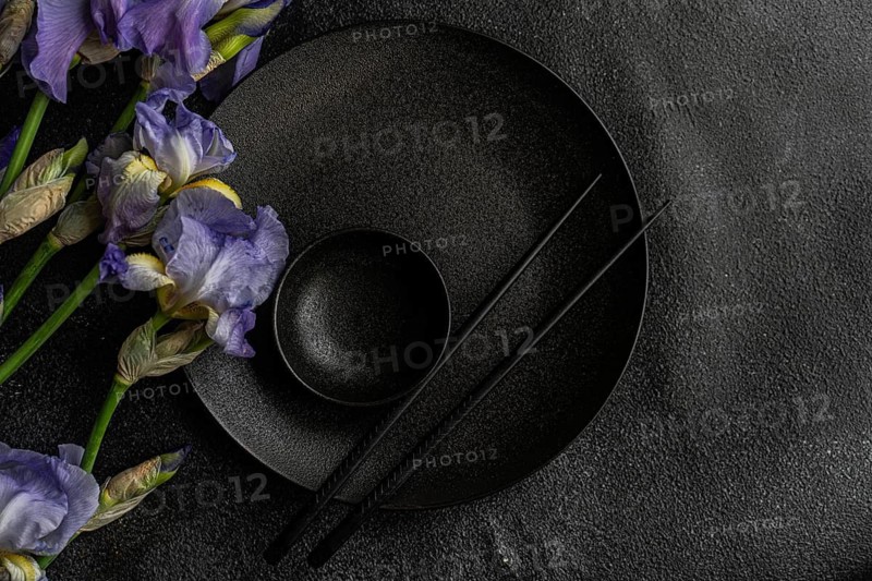 Top view of a sophisticated black dinnerware set complemented by delicate Iris flowers on a dark textured background. Ideal for modern table decor concepts