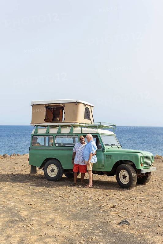 A joyful gay senior couple embraces beside an off-road vehicle during a scenic camping trip by the sea. Both are smiling, dressed in casual summer clothes, with a clear blue sky and calm sea in the background