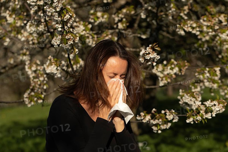 Woman sneezing into a tissue in a blooming orchard during spring. Pollen allergy or hay fever concept