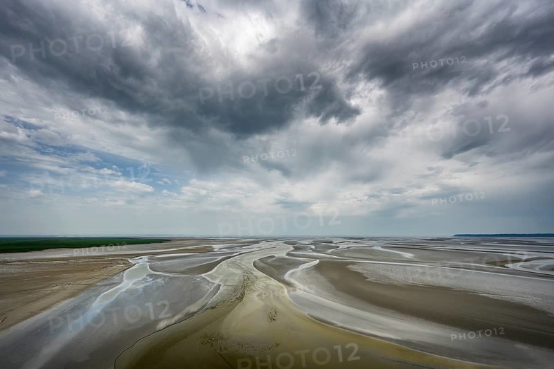 View from the rocky island, monastery island of Le Mont Saint Michel onto a wide mudflat landscape with creeks and dramatic clouds at low tide, Mont-Saint-Michel Bay, Pontorson, Manche department, Normandy, France