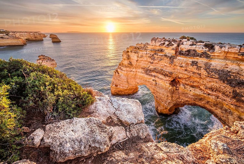 Sunrise view of Praia da Marinha beach in Algarve, Portugal, with a limestone sea arch, Atlantic water, warm sky, and rugged coastal cliffs
