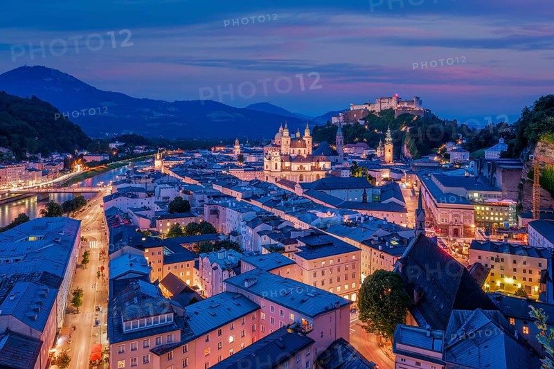 A wide aerial perspective of Salzburg, Austria, featuring the Salzach river flowing past the illuminated historic center and Hohensalzburg Fortress at twilight