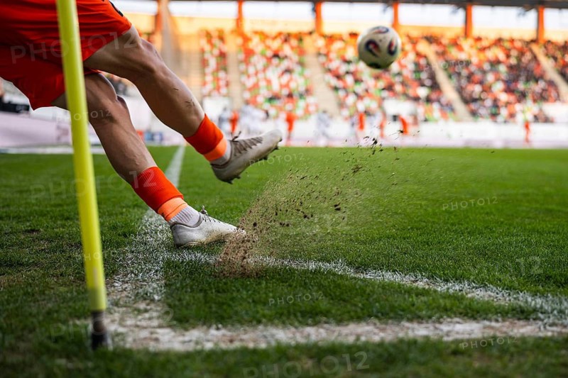 A close-up of a soccer player's legs kicking a ball during a corner kick with turf and dust flying up at a stadium in Lubin, Poland