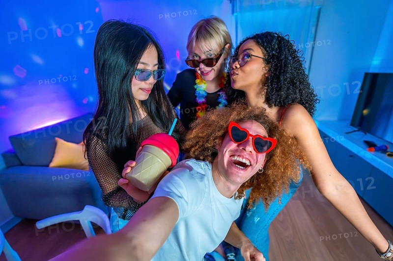 Group of cheerful multi ethnic friends taking a funny selfie at a neon lit home party, enjoying drinks and having fun