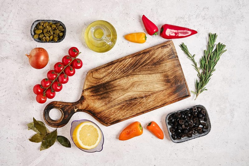 Colorful vegetables and fresh herbs are laid out around wooden cutting board. Cherry tomatoes, bell peppers, olives, and olive oil are ready for cooking