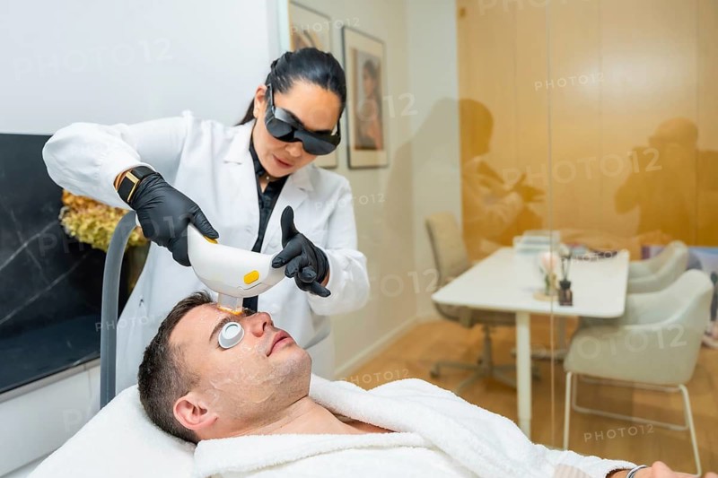 Man undergoing a non invasive laser facial treatment for skin rejuvenation and care, performed by a female doctor wearing protective glasses in a modern aesthetic clinic