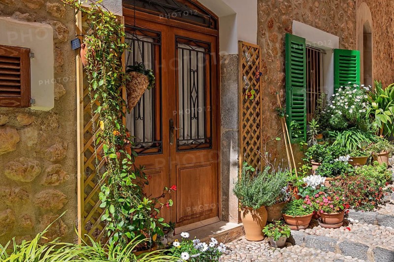Charming stone house entrance with a wooden door surrounded by flower pots and climbing plants with hanging flower baskets in a alley, Fornalutx, Mallorca, Spain