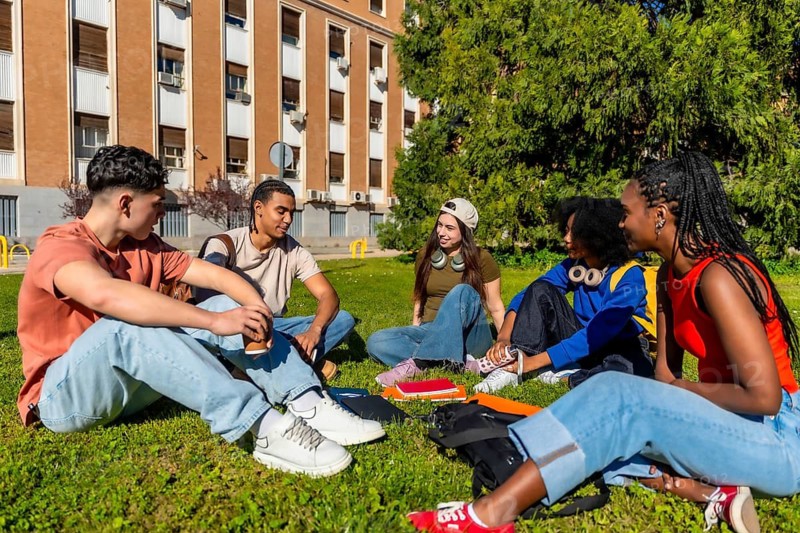 Diverse university students sitting on green campus grass, chatting, smiling and relaxing together between classes on a sunny day, enjoying friendship and study break