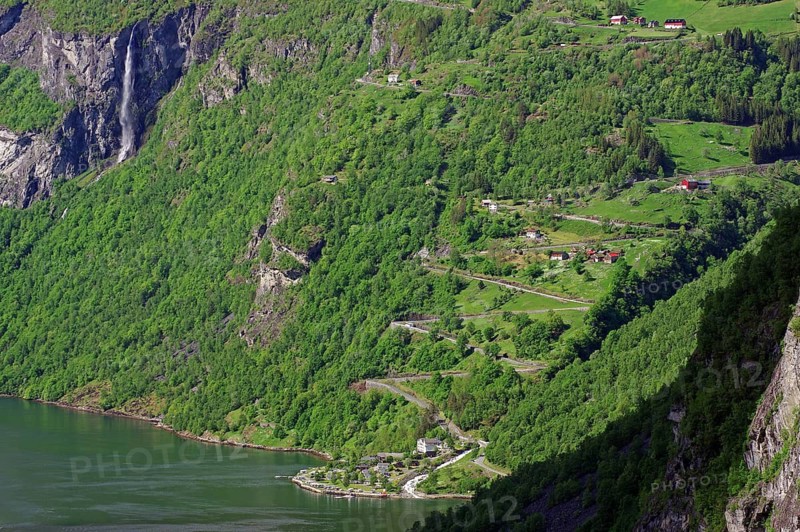 A green, mountainous landscape with a winding road leading up a zig-zag mountain, a few small houses and a waterfall that flows into a river, Eagle Road, Geiranger, Geirangerfjord, Norway