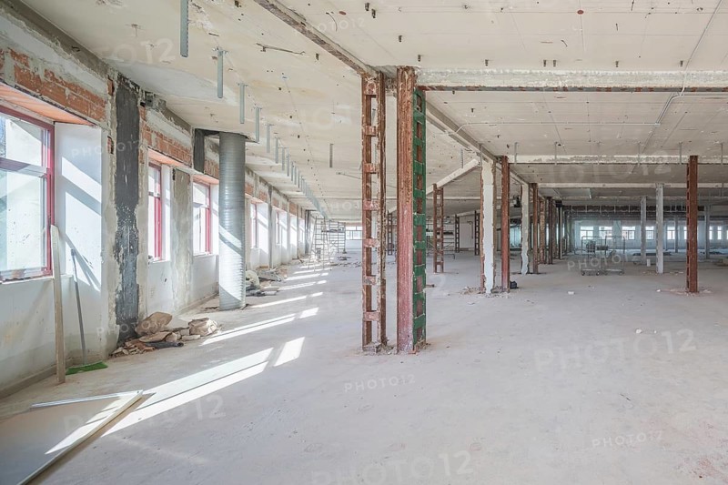 A large, empty industrial space with exposed beams and scattered construction materials. Sunlight enters through the windows, highlighting the raw and unfinished elements