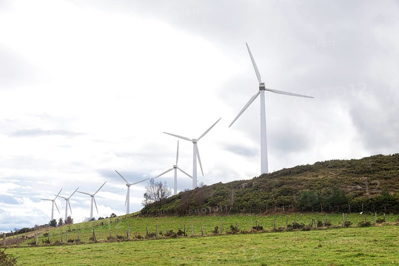 A row of windmills lines a lush, green hillside, harnessing wind energy beneath a cloudy sky. The scene captures the blend of technology and nature, promoting renewable energy