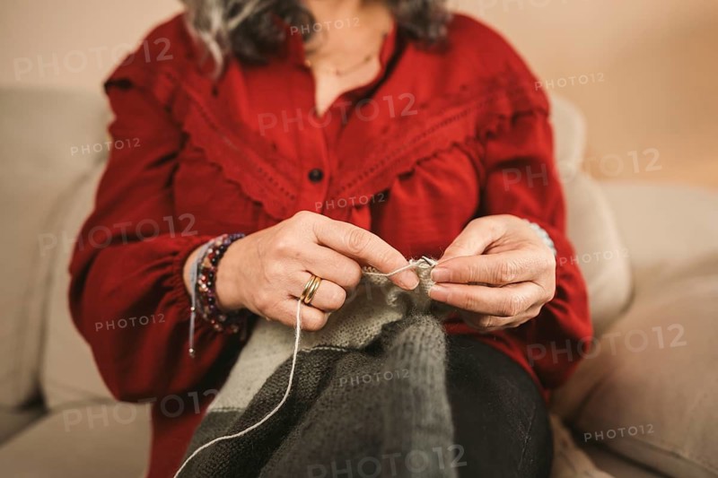 A woman in a red blouse skillfully knits a gray fabric, her fingers gracefully maneuvering the yarn. The scene captures the calm and focused essence of a traditional craft