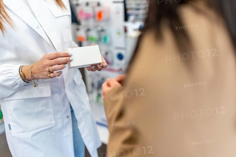 Pharmacist in white coat holding white medicine box, explaining prescription and dispensing medication to female client, highlighting healthcare and assistance service in drugstore