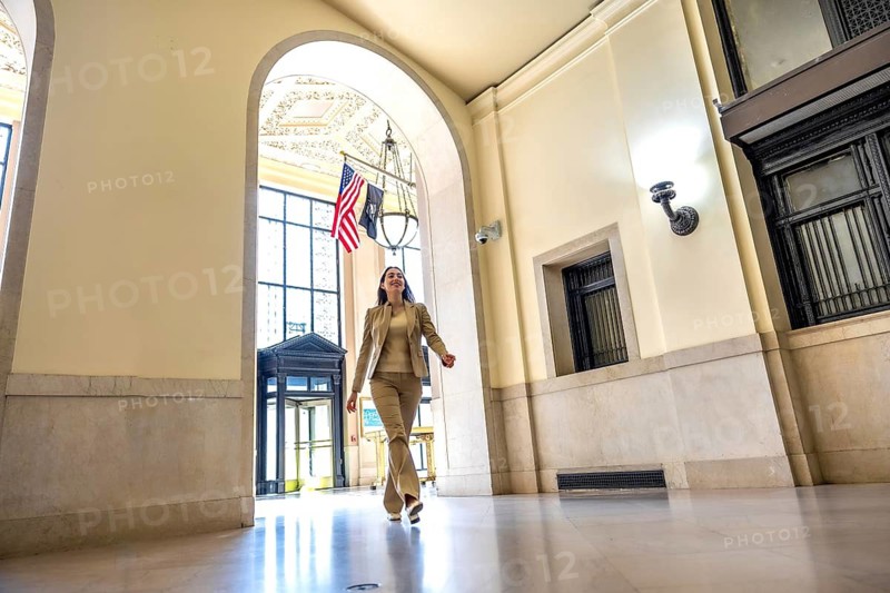 Professional woman in a business suit confidently walking through a grand archway inside a historic government building lobby, featuring classic architecture and an american flag