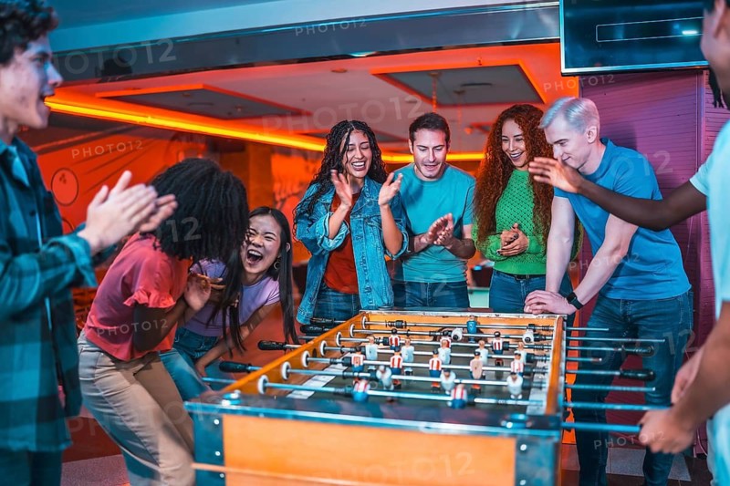 Multiethnic friends enjoying a foosball game, clapping and laughing, inside a vibrant entertainment venue
