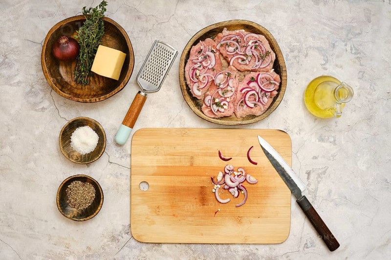 Fresh pork chops are arranged on a wooden plate with sliced onions and herbs nearby. A cutting board holds finely chopped onions, while olive oil and cheese are ready for cooking