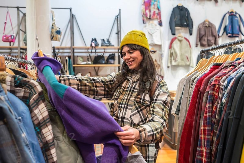 Young woman smiling as she browses a colorful rack of pre loved clothing in a thrift shop, enjoying sustainable, budget friendly fashion and eco conscious shopping in store