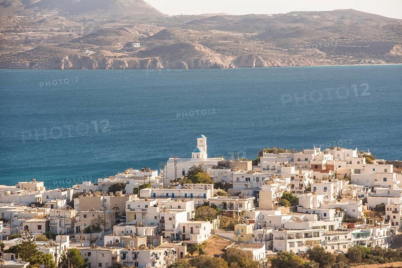 Aerial view of Adamas village on Milos Island, Greece. The scene captures white buildings, a charming church, and the deep blue Aegean Sea, creating a stunning landscape