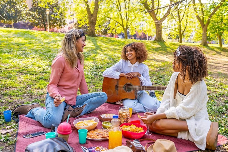 Three female friends having fun while picnicking on a blanket in a park, eating snacks, drinking beverages, and playing music