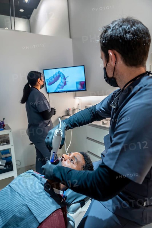 Dentist using intraoral scanner on patient's teeth, with dental assistant and 3d model displayed on screen in background