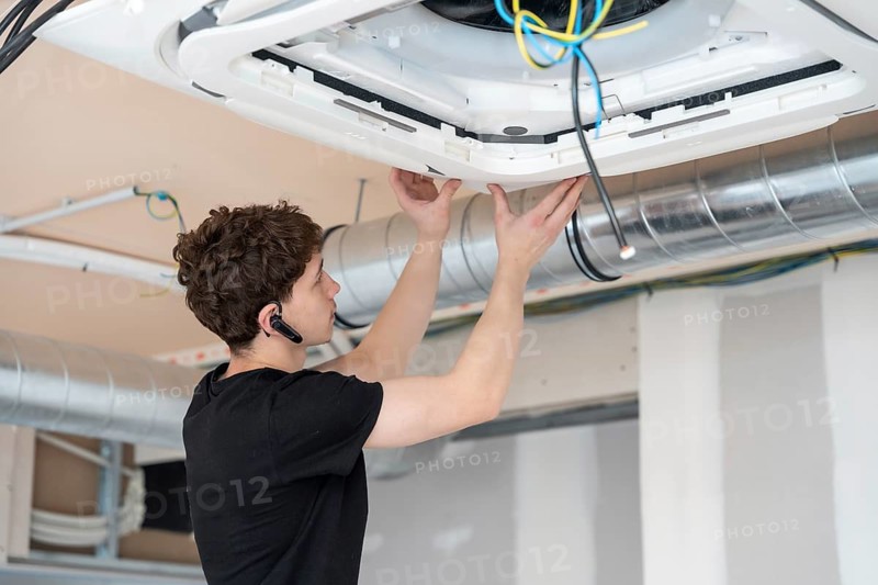 Young male technician installing a modern cassette air conditioning unit into a ceiling during commercial or residential construction, connecting wires and adjusting the panel