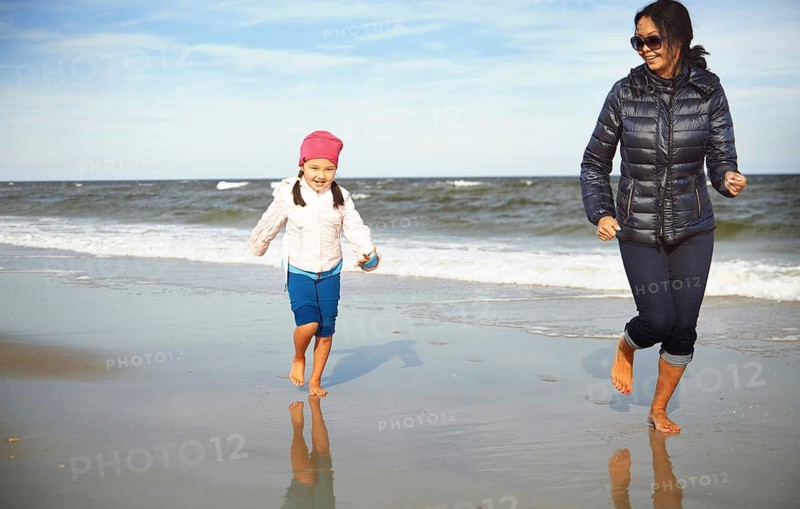 A joyful scene featuring a mother and daughter running barefoot on a sandy beach in California, United States. The ocean waves and clear sky create a lively and refreshing atmosphere
