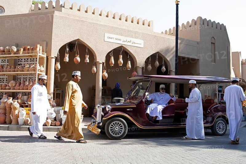 Omani men in traditional dress and a tourist vehicle in the souk or soug or market in the old town of Nizwa, ad-Dachiliyya region, Oman