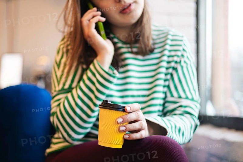 A cropped unrecognizable woman enjoys a coffee break in a cafe, holding a disposable cup and talking on a green smartphone, closely cropped to focus on her hands and accessories
