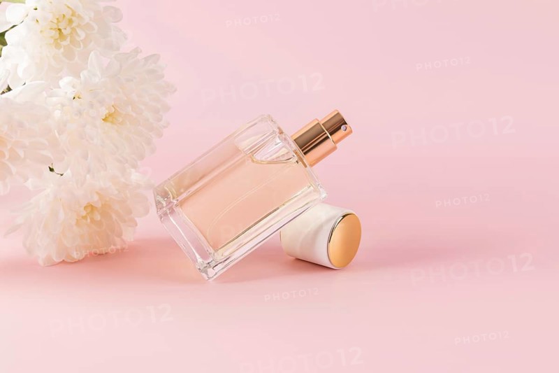A delicate glass perfume bottle lies elegantly next to soft white flowers on a pastel pink background. The scene exudes sophistication and serenity, ideal for luxury branding