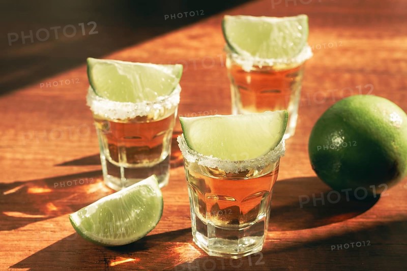 Three glasses with tequila and salt on the rims are arranged on a wooden table. Each glass has a slice of lime on top. A lime sits in the background under natural light