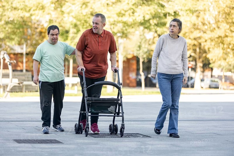 Three individuals with intellectual disabilities walk together. One man uses a mobility walker while another offers support, highlighting inclusion, independence, and companionship in daily life