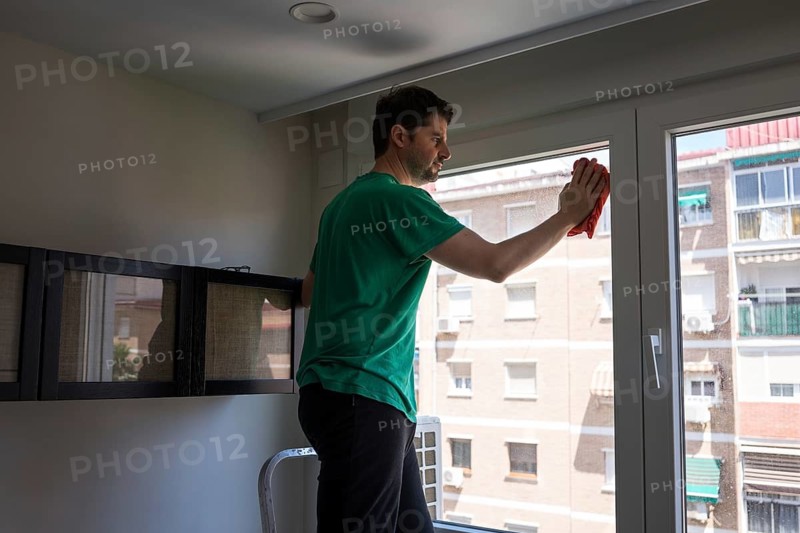 A man performs household chores in an urban apartment, focusing on cleaning the window. The image captures the essence of everyday life and maintenance in a modern home