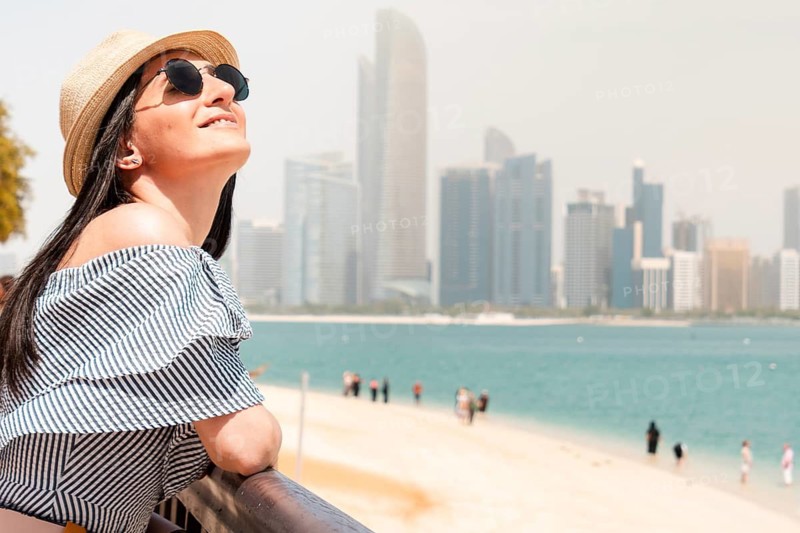 Happy young female tourist on viewpoint in hot day visiting Abu dhabi, UAE - Traveler girl taking portrait in front of capital skyline background. Urban middle east city life travel attractions