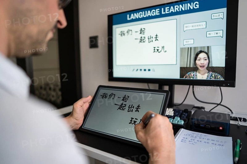 A man learning Mandarin online at home, engaging with a virtual instructor via video call. He writes Chinese characters on a digital tablet, immersed in the language lesson