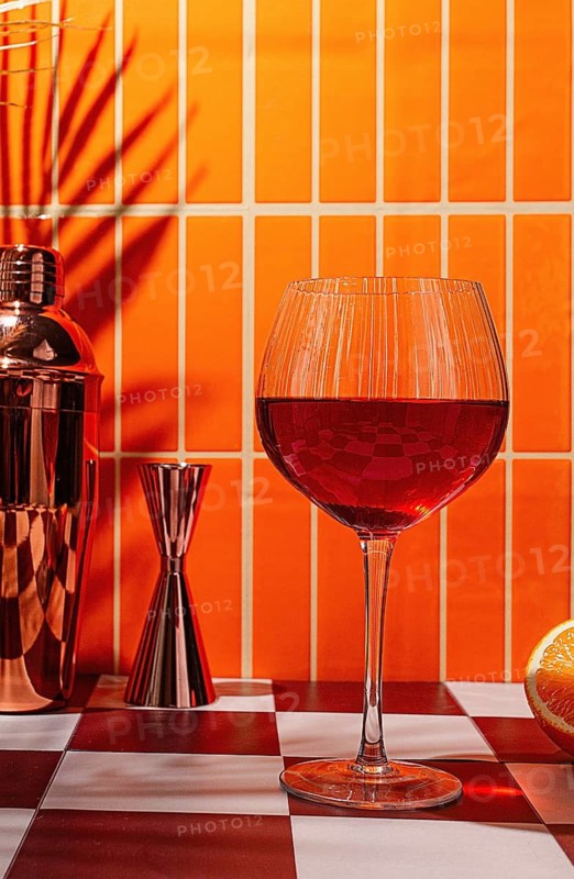 A cocktail glass with a red drink stands on a checkered table. Nearby, a cocktail shaker and a jigger are visible. Bright orange tiles create an eye-catching backdrop
