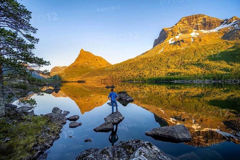 Young man in front of picturesque mountain landscape in the Innerdalen high valley with Innerdalstårnet summit, mountain peaks reflected in lake Innerdalsvatna, in the evening light at sunset, Trollheimen Mountain Area, Sunndal, Møre og Romsdal, Norway