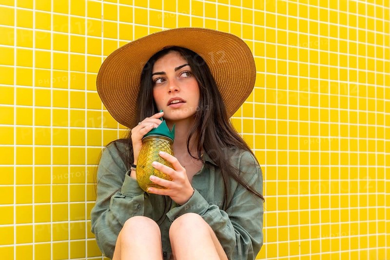 Woman wearing a straw hat and green shirt, taking a sip from a pineapple shaped jar while sitting casually against a vibrant yellow tiled wall on a sunny summer day