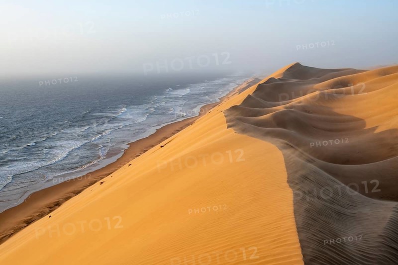 Sand dunes and sea in evening light, wind blowing sand across the dunes, view of the Atlantic coast from high sand dunes, impressive desert landscape at sunset, Sandwich Harbour, Namib Naukluft Park, Namibia