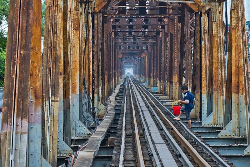 A Vietnamese State Railroad train crosses the Long Bien Railway Bridge across the Red River while a man crosses the tracks with a red bucket, Hanoi, Ha Noi province, Vietnam