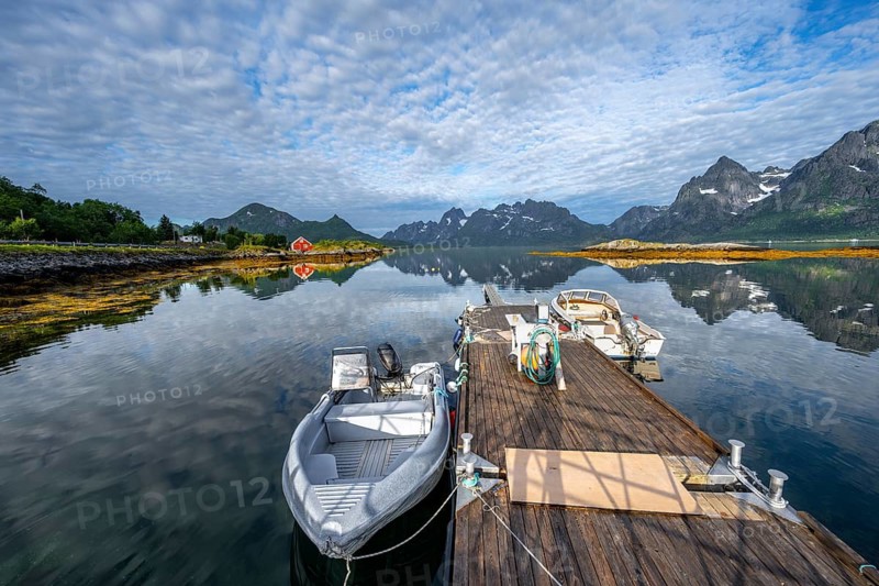 Red fishing hut on the shore and boats on a jetty, Rorbuer hut, picturesque fjord landscape at Raftsund fjord, mountains reflected in the water, Vesterålen, Norway