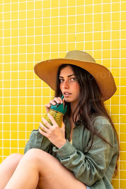 Young woman sitting by a bright yellow tiled wall, enjoying a cool drink from a pineapple shaped bottle, embodying carefree summer leisure and youthful relaxation