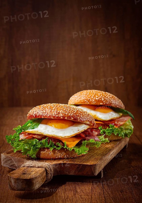Two juicy burgers sit on a wooden board, each filled with lettuce, bacon, cheese, and fried eggs
