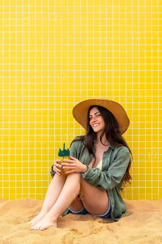 Young woman sits on warm sand against a yellow tiled wall, smiling and holding a pineapple shaped tropical drink, evoking summer relaxation, joy, and carefree vacation vibes