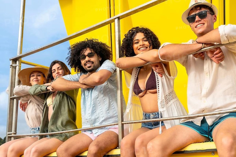 Diverse group of young adult friends smiling and relaxing while sitting on a bright yellow structure during their summer vacation, symbolizing happiness, youth, and travel