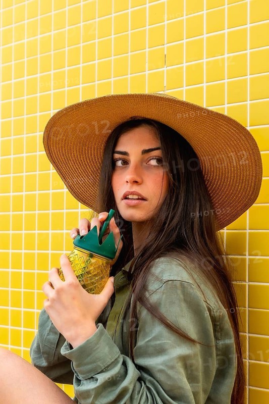 Young woman wearing a straw hat and green shirt, sitting against a bright yellow tiled wall, holding a pineapple shaped cup and drinking, embodying a vibrant summer lifestyle
